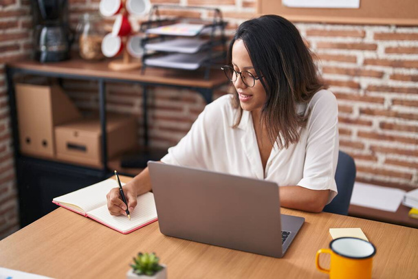 Młoda latynoska kobieta biznes pracownik za pomocą laptopa pisanie na notatniku w biurze - Zdjęcie, obraz