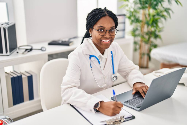 African american woman doctor using laptop writing medical report at clinic - Foto, afbeelding