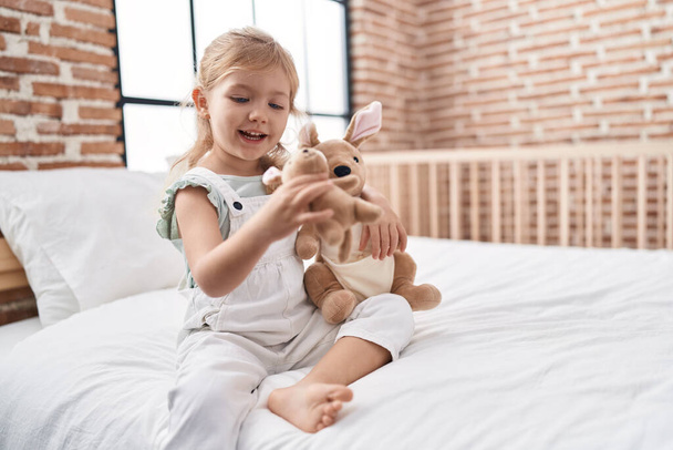 Adorable chica rubia jugando con la muñeca sentada en la cama en el dormitorio - Foto, Imagen
