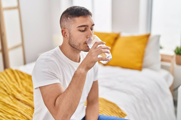 Jovem hispânico bebendo copo de água sentado na cama no quarto - Foto, Imagem