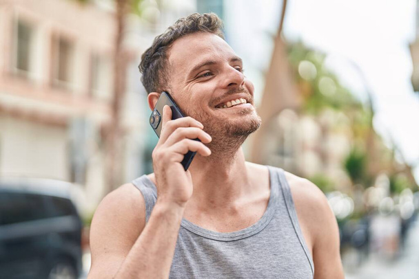 Νεαρός Ισπανόφωνος άντρας χαμογελάει με αυτοπεποίθηση μιλώντας στο smartphone στο δρόμο - Φωτογραφία, εικόνα