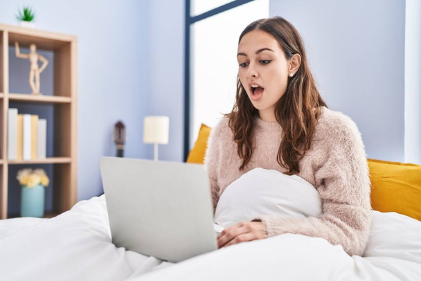 Mujer hispana joven usando computadora portátil en la cama asustada y sorprendida con la boca abierta para sorpresa, cara de incredulidad  - Foto, Imagen