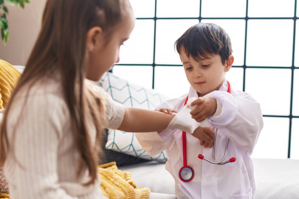 Adorable boy and girl wearing doctor uniform bandaging arm at home - Zdjęcie, obraz