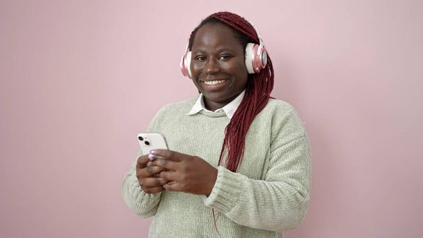 Αφρικανική γυναίκα με πλεγμένα μαλλιά χρησιμοποιώντας smartphone φορώντας ακουστικά πάνω από απομονωμένο ροζ φόντο - Φωτογραφία, εικόνα