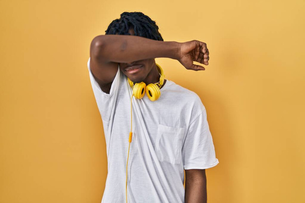 Giovane uomo africano con dreadlocks in piedi su sfondo giallo che copre gli occhi con il braccio, guardando serio e triste. concetto di avvistamento, nascondiglio e rifiuto  - Foto, immagini