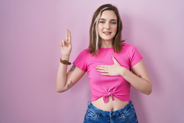 Blonde caucasian woman standing over pink background smiling swearing with hand on chest and fingers up, making a loyalty promise oath  - Φωτογραφία, εικόνα