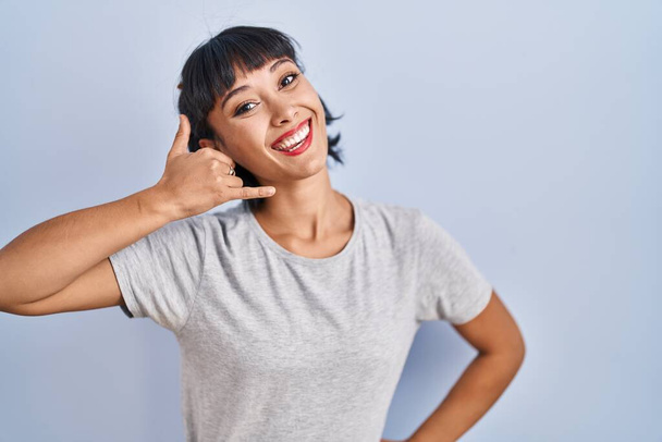青い背景の上にカジュアルなTシャツを着た若いヒスパニック系の女性は、電話で話すような手や指で電話のジェスチャーをして笑っています。コミュニケーションの概念.  - 写真・画像