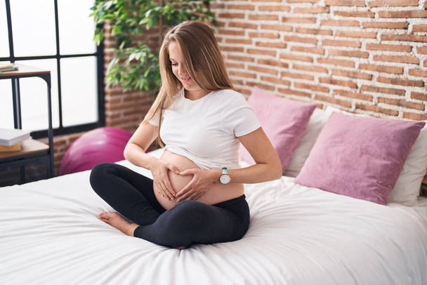 Fiatal terhes nő mosolygós magabiztos csinál szív gesztus hason a hálószobában - Fotó, kép
