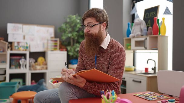 Young redhead man preschool teacher writing on notebook sitting on chair at kindergarten - Foto, imagen