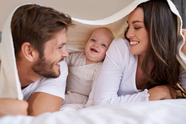 Ευτυχισμένη μητέρα, πατέρας και μωρό με κουβέρτα στο κρεβάτι για αγάπη, φροντίδα και ποιοτικό χρόνο μαζί. Γονείς, οικογένεια και παιχνιδιάρικο νεογέννητο παιδί χαλαρώνουν στο υπνοδωμάτιο με κρεβάτι φρούριο, χαμόγελο και δέσιμο στο σπίτι. - Φωτογραφία, εικόνα