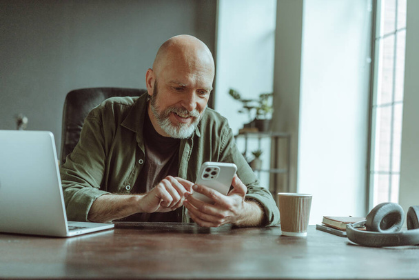 Mature old man comfortably seated home, actively engaged in texting message on phone. Desk adorned with laptop, headphones, and cup of coffee, indicating setup conducive to work and relaxation. . High - Photo, Image