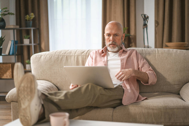 Vanhempi mies, jolla on arvostettu hopeaparta, kuvasi nauttimista kotona. Hän istui mukavasti sohvalla, engrossed katsomassa internetiä hänen laptop. Laadukas kuva - Valokuva, kuva