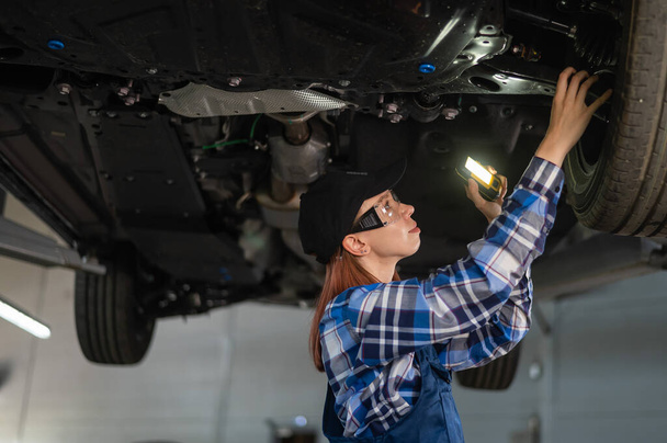 A female mechanic inspects a lifted car. A girl at a mans work - Zdjęcie, obraz