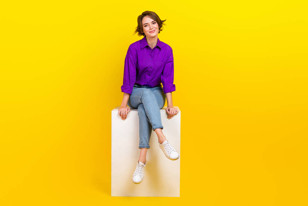 Full size photo of pretty young girl sitting white cube smile wear trendy violet smart casual outfit isolated on yellow color background. - Fotó, kép