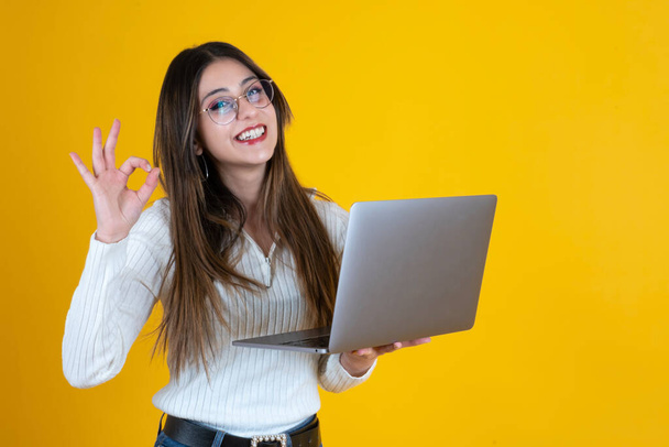 Segurando laptop e gesto ok sinal, sorrindo jovem mulher caucasiana segurando laptop e gesto ok sinal. Trabalhadora de escritório do sexo feminino usando computador e mostrando ok com a mão. Espaço de cópia. - Foto, Imagem
