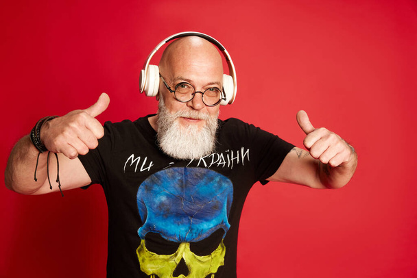 Portrait fp mature bearded, bald man with positive facial expression, listening to music in headphones against red studio background. Concept of human emotions, lifestyle, male fashion. Rock-n-roll - 写真・画像