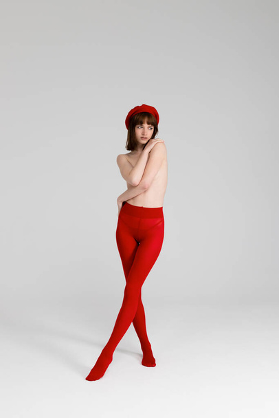 Young beautiful girl only in red tight and a beret, hands covering her bare chest. Sensual slim girl posing topless. Isolated on white background. Full length, vertical. - Photo, Image
