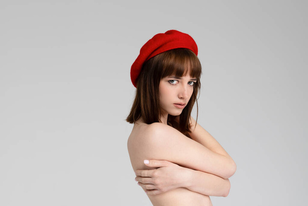 Ένα νεαρό όμορφο κορίτσι μόνο σε κόκκινο σφιχτά και ένα μπερέ, που καλύπτει το γυμνό στήθος της. Αισθησιακό λεπτό κορίτσι που ποζάρει γυμνόστηθη. Απομονωμένο σε λευκό φόντο. Πορτρέτο, φωτογραφία κεφαλής. - Φωτογραφία, εικόνα
