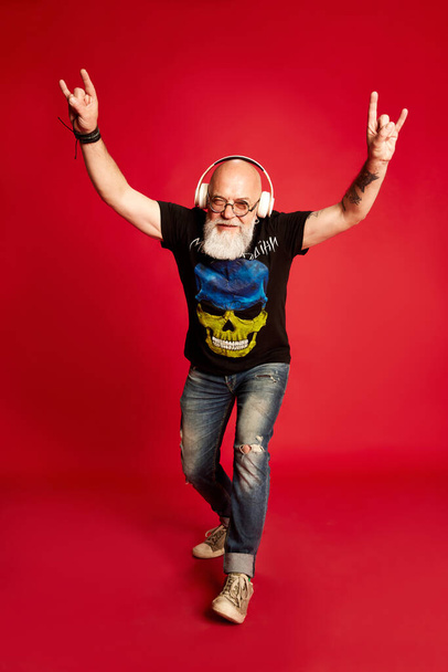 Rock-n-roll. Full-length portrait emotional, mature, bearded man with tattoos, listening to music in headphones against red studio background. Concept of human emotions, lifestyle, male fashion - Zdjęcie, obraz