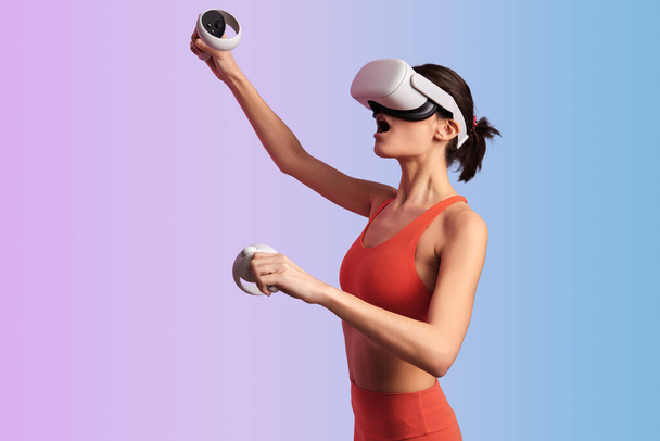 Side view of young female in VR goggles and with controllers playing video game against gradient background - Fotografie, Obrázek