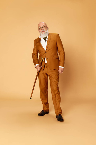 Portrait of bearded, mature, bald man in elegant, classical suit and walking stick posing with serious expression against studio background. Concept of human emotions, lifestyle, fashion, business - Fotoğraf, Görsel