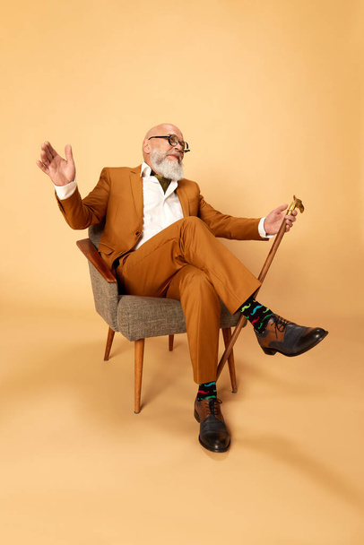Portrait of bearded, mature, bald man in elegant, classical suit and walking stick, sitting in armchair and smiling against studio background. Concept of human emotions, lifestyle, fashion, business - Foto, Imagem
