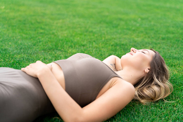 Young Caucasian slim fit woman with closed eyes in stylish fitness clothes lying on green grass loan relaxing, sleeping or dreaming after workout or running on summer weekend. Leisure activity, hobby - Φωτογραφία, εικόνα