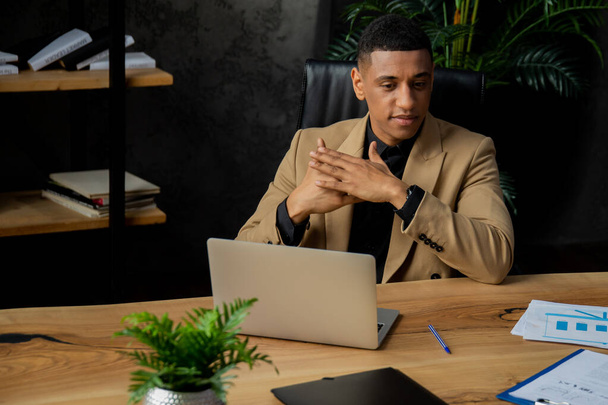 Een Afro-Amerikaanse ondernemer zit aan een bureau in zijn kantoor. Een man in een bruin jasje zit voor een laptop en denkt ergens aan. Hoge kwaliteit foto - Foto, afbeelding