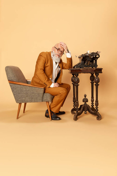 Working crisis. Portrait of bearded, mature, bald man in elegant, classical suit sitting near typewriter and holding hand on head. Ideas. Concept of human emotions, lifestyle, business, art, fashion - Φωτογραφία, εικόνα
