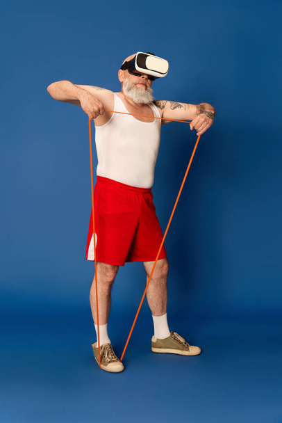 Портрет дорослого бородатого чоловіка в спортивному одязі і віртуальних окулярах тренування з розширювачами фітнесу на синьому студійному фоні. Онлайн-тренінги. Концепція спорту, активний спосіб життя, здоров'я
 - Фото, зображення