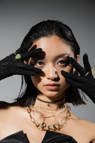 ritratto di giovane donna bruna e asiatica con i capelli corti posa in guanti neri con anelli d'oro, guardando la fotocamera su sfondo grigio, acconciatura bagnata, mani vicino al viso, trucco naturale - Foto, immagini