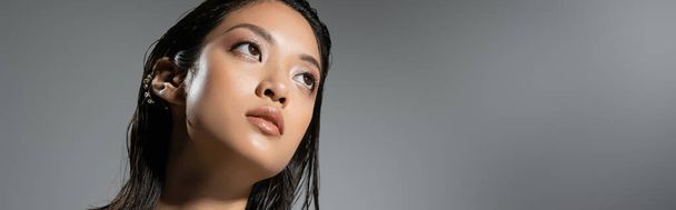portrait of mesmerizing asian young woman with short hair and golden earrings posing while looking away on grey background, wet hairstyle, natural makeup, looking away, banner - Photo, Image