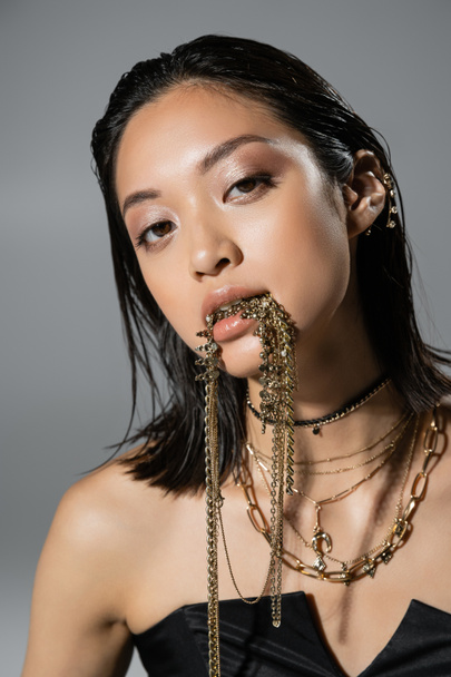 retrato de la mujer joven asiática de moda con pelo corto posando en vestido negro sin tirantes mientras sostiene joyas de oro en la boca sobre fondo gris, peinado húmedo, maquillaje natural, mirando a la cámara - Foto, Imagen