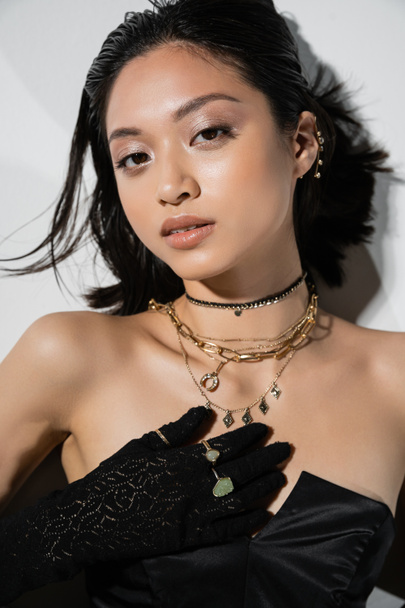 top view of alluring asian young woman with short hair lying in black gloves and strapless dress while posing in golden jewelry on grey background, wet hairstyle, natural makeup, looking at camera - Fotó, kép