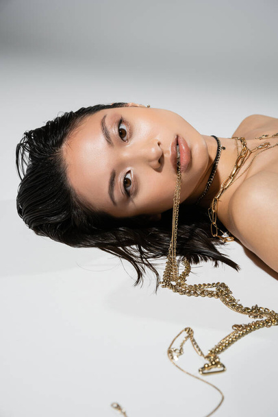 young asian woman with short brunette hair holding golden jewelry in mouth while looking at camera and lying on grey background, everyday makeup, wet hairstyle, brown eyes - Foto, Imagem