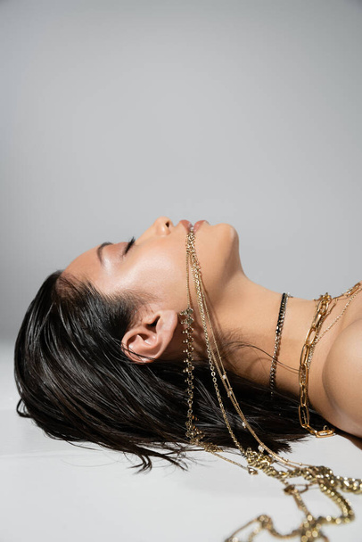vista lateral de mujer asiática joven con pelo corto morena sosteniendo joyas de oro en la boca mientras está acostado sobre fondo gris, maquillaje diario, peinado mojado, ojos cerrados  - Foto, Imagen