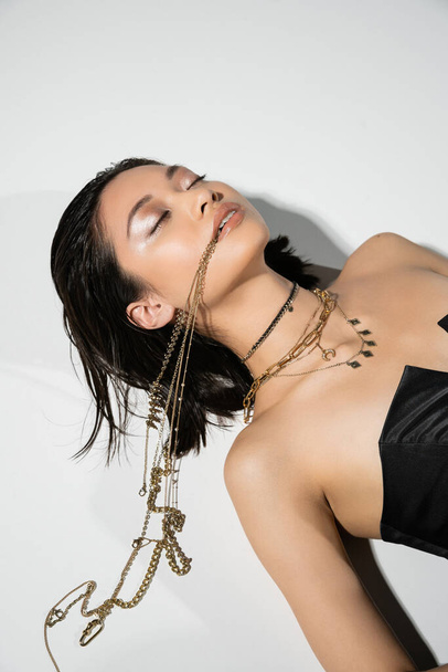 high angle view of young asian woman with short brunette hair holding golden jewelry in mouth while lying on grey background, everyday makeup, wet hairstyle, closed eyes  - Foto, immagini