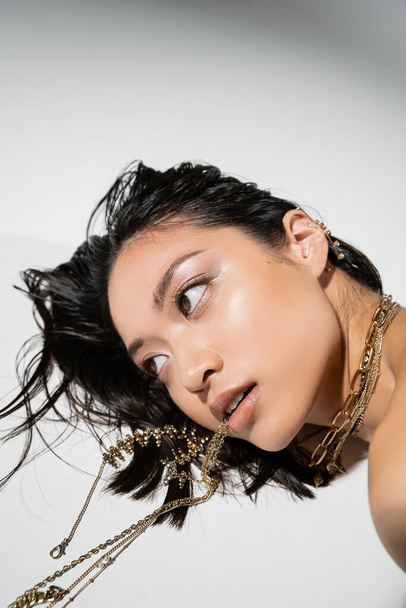 stunning asian model with short brunette hair holding golden jewelry in mouth while lying on grey background, everyday makeup, wet hairstyle, young woman, looking away - Φωτογραφία, εικόνα