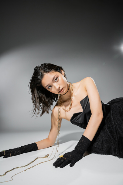 asian model with short brunette hair holding golden jewelry in mouth while looking at camera and posing in black strapless dress on grey background, everyday makeup, wet hairstyle, young woman  - Photo, Image