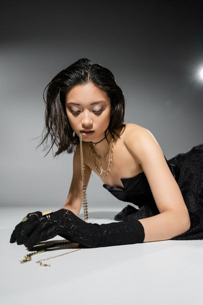 podmanivý asijský model s krátkými brunetkami vlasy drží zlaté šperky v ústech při pohledu dolů a pózování v bezramkové šaty na šedém pozadí, každodenní make-up, mokré vlasy, mladá žena  - Fotografie, Obrázek