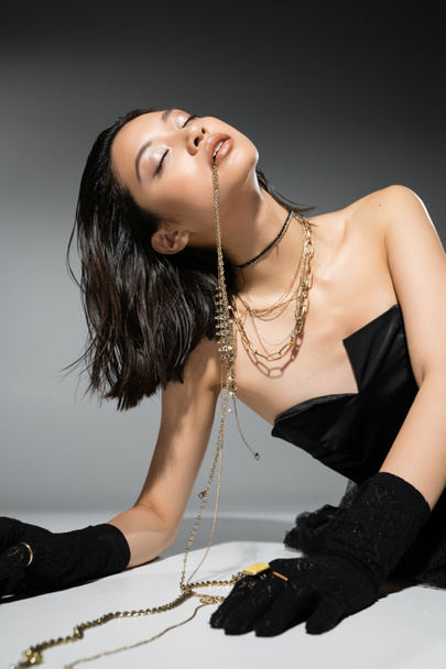 okouzlující asijský model s krátkými brunetkami vlasy drží zlaté šperky v ústech, zatímco pózuje v bezramkové šaty na šedém pozadí, každodenní make-up, mokrý účes, mladá žena, zavřené oči - Fotografie, Obrázek