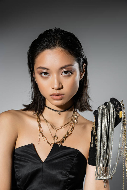 portrait of brunette and asian young woman with short hair holding golden and silver jewelry while wearing glove and standing in strapless dress on grey background, wet hairstyle, natural makeup - Foto, afbeelding