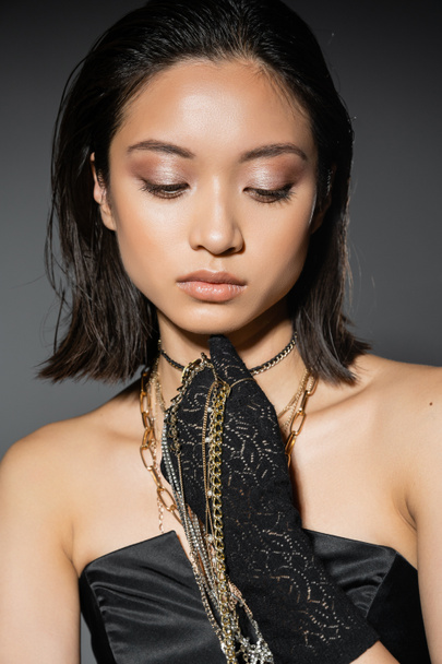 portrait of alluring asian young woman with short hair holding golden and silver jewelry while wearing glove and standing in strapless dress grey background, wet hairstyle, natural makeup - Photo, Image