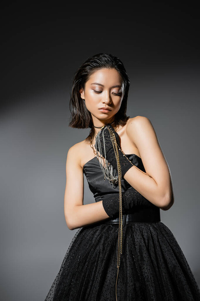 charming asian young woman with short hair holding golden and silver jewelry while wearing gloves and standing in elegant strapless dress on grey background, wet hairstyle, natural makeup - Photo, Image