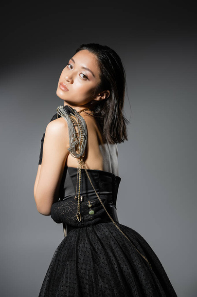 portrait of brunette and asian young woman with short hair posing with golden and silver jewelry on shoulder while standing in black strapless dress on grey background, wet hairstyle, natural makeup - Photo, Image