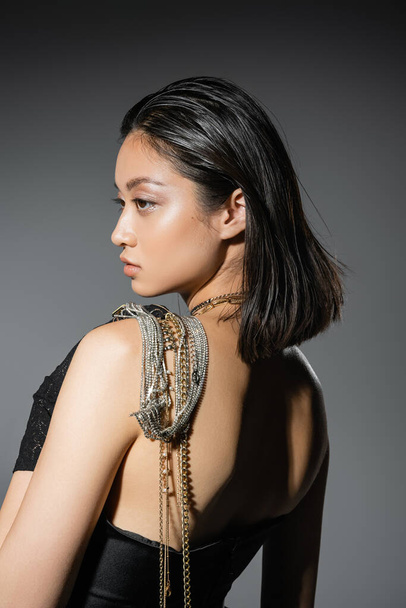 portrait of brunette and asian young woman with short hair posing with golden and silver jewelry on shoulder while standing in strapless dress on grey background, wet hairstyle, natural makeup, side view - Fotografie, Obrázek