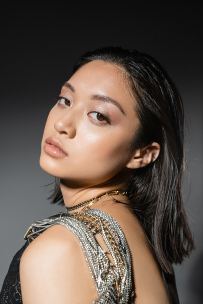 portrait of brunette and asian young woman with short hair posing with golden and silver jewelry on shoulder and looking at camera on grey background, wet hairstyle, natural makeup - Foto, imagen