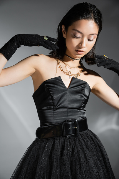 glamorous asian young woman with short hair posing in black strapless dress with belt and gloves while looking down on grey background, wet hairstyle, golden necklaces  - Zdjęcie, obraz
