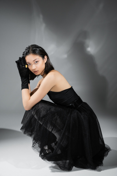full length of mesmerizing asian young woman with short hair sitting in black strapless dress with tulle skirt and gloves while looking at camera on grey background, wet hairstyle, golden necklaces  - Fotografie, Obrázek