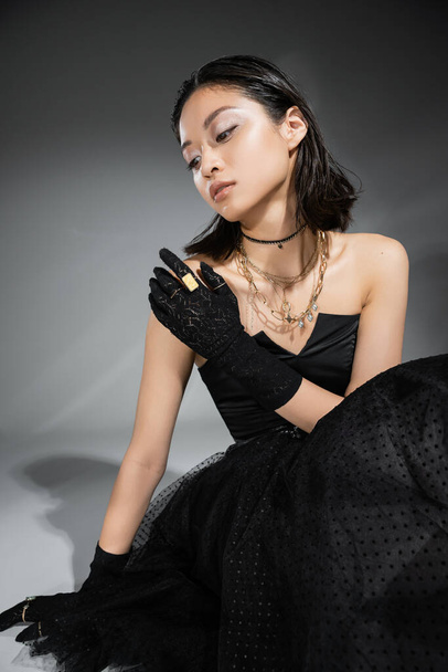charming asian young woman with short hair sitting in black strapless dress with tulle skirt and gloves while looking away on grey background, wet hairstyle, golden necklaces and rings  - Фото, изображение